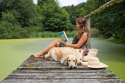 Junge Frau sitzt auf Steg am Teich und liest ein Buch während Golden Retriever Moana geduldig wartet, Haunetal, Rhön, Hessen, Deutschland [MR]