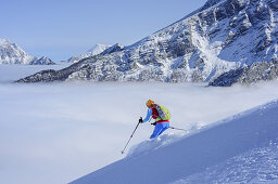 Woman back-country skiing downhill from Hochalm, Watzmann and sea of fog in background, Hochalm, Hochkalter, National Park Berchtesgaden, Berchtesgaden Alps, Berchtesgaden, Upper Bavaria, Bavaria, Germany