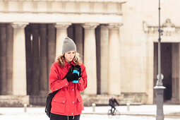 Junge Frau mit Isolierbecher am verschneiten Königsplatz in München, Bayern, Deutschland