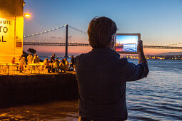 tourist with Tablet photographs sunset on riverfront, restaurant Ponto Final, view from south bank of River Tagus,and the 25th April Bridge, Cacilhas, Almada, Lisbon, Portugal