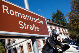 Motorcycle rider behind road sign for Romantische Strasse romantic road through Unteres Taubertal, Bronnbach, near Wertheim, Spessart-Mainland, Franconia, Baden-Wuerttemberg, Germany