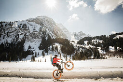 Ein junger Mann zeigt ein Wheelie auf einem Fatbike, Snowbike, Mountainbike vor dem zugefrorenen Lauenensee (Louwenesee) in der Nähe von Gstaad, Berner Oberland, Schweiz