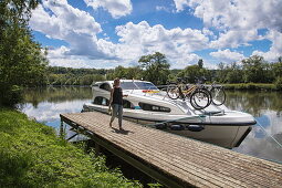 Junge Frau läuft entlang Steg und trägt Weißwein vor Le Boat Elegance Hausboot an Anlegestelle während Bootstörn auf Fluss Petit Saône, Soing, Soing-Cubry-Charentenay, Haute-Saône, Bourgogne Franche-Comté (Burgund), Frankreich, Europa