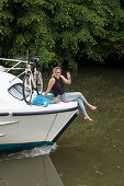 Junge Frau sitzt am Bug von Le Boat Magnifique Hausboot und erhebt Weinglas während Bootstörn auf Fluss Petit Saône, nahe Soing, Soing-Cubry-Charentenay, Haute-Saône, Bourgogne Franche-Comté (Burgund), Frankreich, Europa