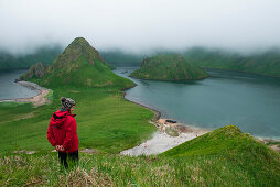 Ein Tourist in rotem Parka steht in hohem Gras oberhalb der wassergefüllten Kaldera, Yankicha-Insel, Uschischir Inselgruppe, Kurilen, Ochotskisches Meer, Russland, Asien