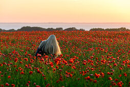 Young woman with long blond hair sits in poppy field on Rügen, Baltic Sea Coast, Mecklenburg-Vorpommern, Germany
