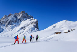 Mehrere Personen auf Skitour gehen auf Almhütten zu, Geislergruppe im Hintergrund, Medalges, Naturpark Puez-Geisler, UNESCO Weltnaturerbe Dolomiten, Dolomiten, Südtirol, Italien