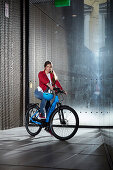 junge Frau auf eBike vor moderner Glasfassade, München, Bayern, Deutschland