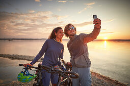 Young  woman on touring bike, young man on touring eBike on tour,taking selfie on  lakeshore, Muensing, bavaria, germany