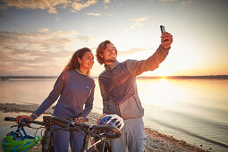 junge Frau auf Tourenrad und junger Mann auf eTourenfahrrad, machen ein selfie am Kiesstrand, Radtour am See, Münsing, Bayern, Deutschland