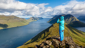 Junge Frau blickt mit dem Fernglas von einem Berg über das Meer und die Fjordlandschaft bei blauem Himmel, Klaksvík, Faröer Inseln