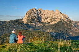 A man and a woman sitting on meadow and views of the Wilder Kaiser mountain, in the dawn, from behind Wilder Kaiser, Kaiser mountains, Tyrol, Austria
