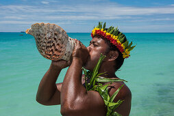 Ein Mann mit Blumen- und Blattkrone bläst in eine große Muschel, um die Ankunft von Besuchern eines Expeditions-Kreuzfahrtschiffes anzukündigen, Butaritari Atoll, Gilbert-Inseln, Kiribati, Südpazifik