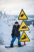 Kleiner Junge auf seinem Snowboard neben Warnschildern, Winter in Finnland
