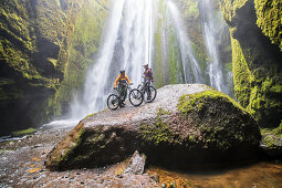 Zwei Mountainbiker in einer Höhle vor einem Wasserfall, Island