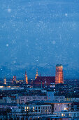 Skyline der Stadt München mit beleuchteter Frauenkirche bei Schneefall bei Nacht \n