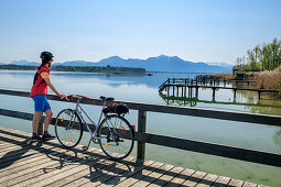 Frau beim Radfahren steht auf Steg und blickt auf Chiemsee, Hochgern im Hintergrund, Chiemseeradweg, Chiemgau, Oberbayern, Bayern, Deutschland