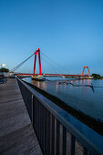 Illuminated, red Willemsbrücke during the blue hour, with plank path and seating on the river bank. Rotterdam, The Netherlands, June 2020