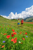 Mann und Frau wandern durch blühendes Mohnfeld mit Castelluccio im Hintergrund, Castelluccio, Sibillinische Berge, Monti Sibillini, Nationalpark Monti Sibillini, Parco nazionale dei Monti Sibillini, Apennin, Marken, Umbrien, Italien