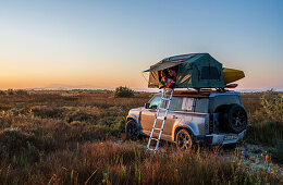 Albania, Southern Europe, young man looks out of the roof tent on an off-road vehicle in the evening sun