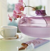 Branch of purple orchids in bulbous glass vase with cup on table