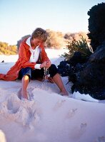 Woman wearing capri jeans and orange Red knee length parka sitting in sand