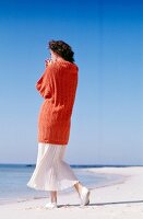 Woman wearing orange turtleneck sweater and airy skirt walking on beach
