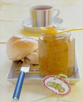 Gooseberry and juniper jam in open jar with bread roll and coffee on plate
