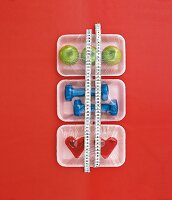 Apples, dumbbells and cakes wrapped in plastic and measure tap on red background