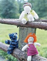 Drei Strickfiguren: zwei Teddybären, eine Puppe auf einem Holzzaun