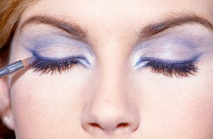 Close-up of woman wearing eye shadow applying blue eyeliner on upper eyelid with brush