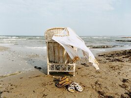 langes weißes Hemd hängt am Korbstuhl am Meer, davor Sandalen + Hut