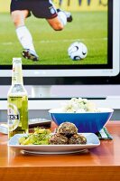 Meatball on plate, potato salad in bowl and beer on wooden table against TV