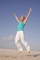 Pretty woman in blue top and white pants jumping on beach with arms outstretched, smiling