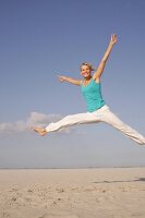 Pretty woman in blue top and white pants jumping on beach with arms outstretched, smiling
