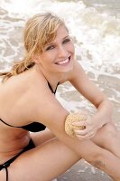 Pretty woman in black bikini rubbing her arm with sponge while sitting on beach, smiling