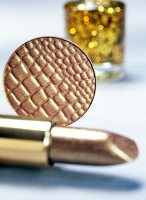 Close-up of gold cosmetic products, lipstick, eye shadow with nail polish in background