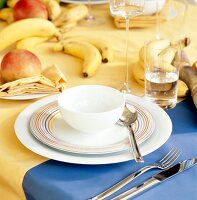 Müslischale auf 2 Tellern, gelbe u. blaue Tischdecken, Bananen, Äpfel.