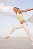 Happy woman in yellow top and white pants holding fluttering cloth and jumping on beach