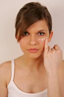 Portrait of pretty woman with brown hair applying cream below her eye with finger