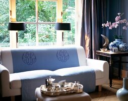Weißes Sofa, blaue Decke mit Initi- alen, Fenster, Lampen