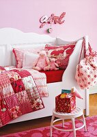 White, wooden, child's bed with scatter cushions and patchwork blankets in shades of red against pink wall