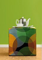 Teekanne und Tasse auf buntem Sitzhocker mit geometrischem Muster im Retro-Stil vor grüner Wand