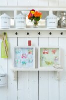 Old storage jars on a white kitchen shelf and framed paper butterflies on a white wooden wall