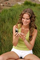 Blonde woman dressed casually and using mobile phone while sitting on grass, smiling