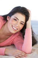 Portrait of pretty woman wearing pink sweater lying on sand with hand on head, smiling