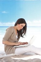 Side view of pretty woman wearing brown cardigan working on laptop while sitting on beach