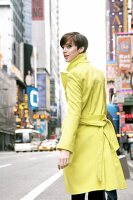 Pretty woman with short hair in yellow overcoat walking on street, looking over shoulder