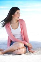 Beautiful woman wearing pink cardigan sitting on beach and looking away, smiling