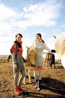 Two women wearing knitwear standing with icelandic horses in field, smiling